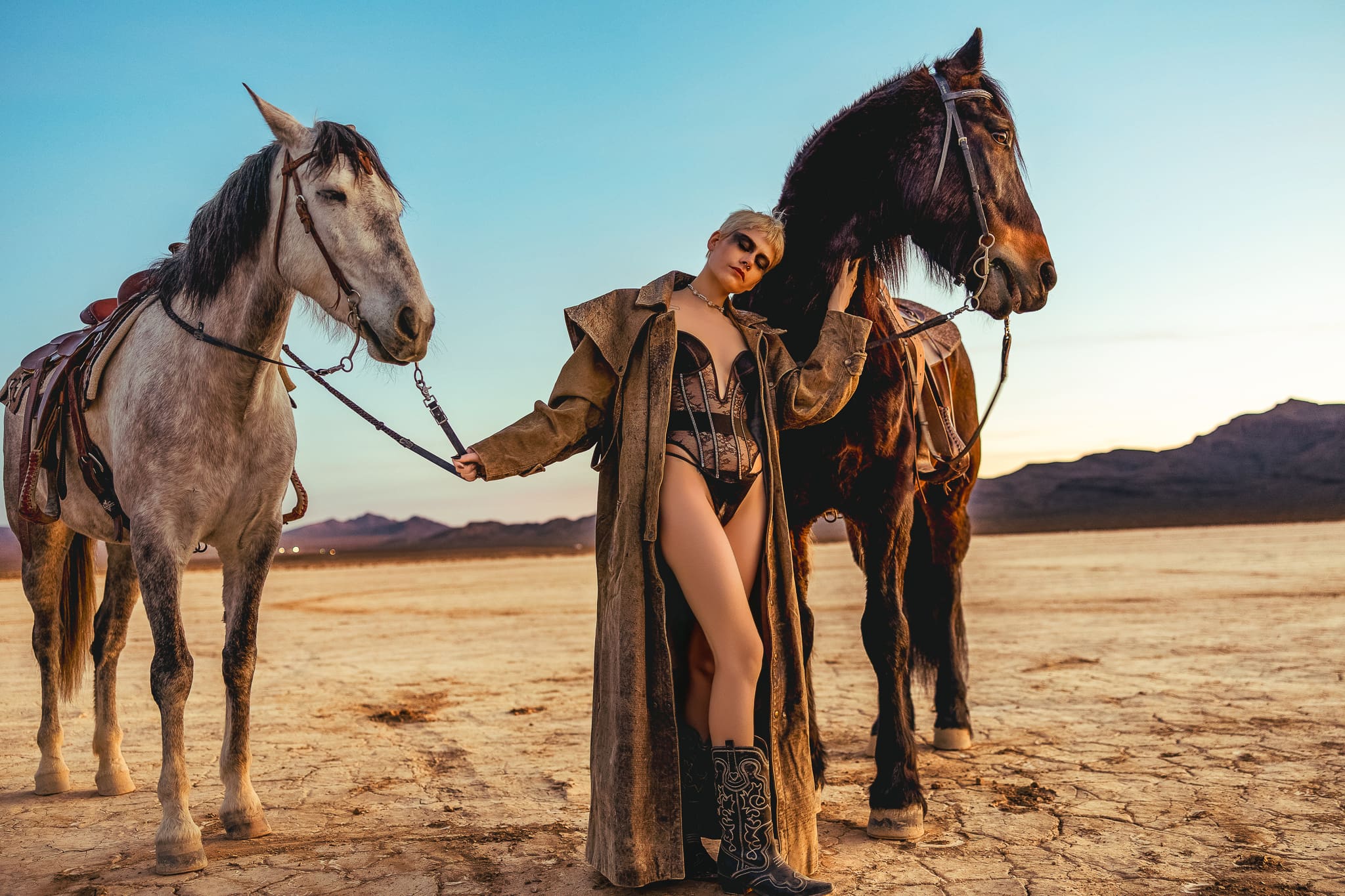 Outdoor boudoir portrait with a woman in lingerie, cowboy boots and a long coat posing with two horses in the desert by Hood River, Oregon boudoir photographer Tobi Rose Photography.