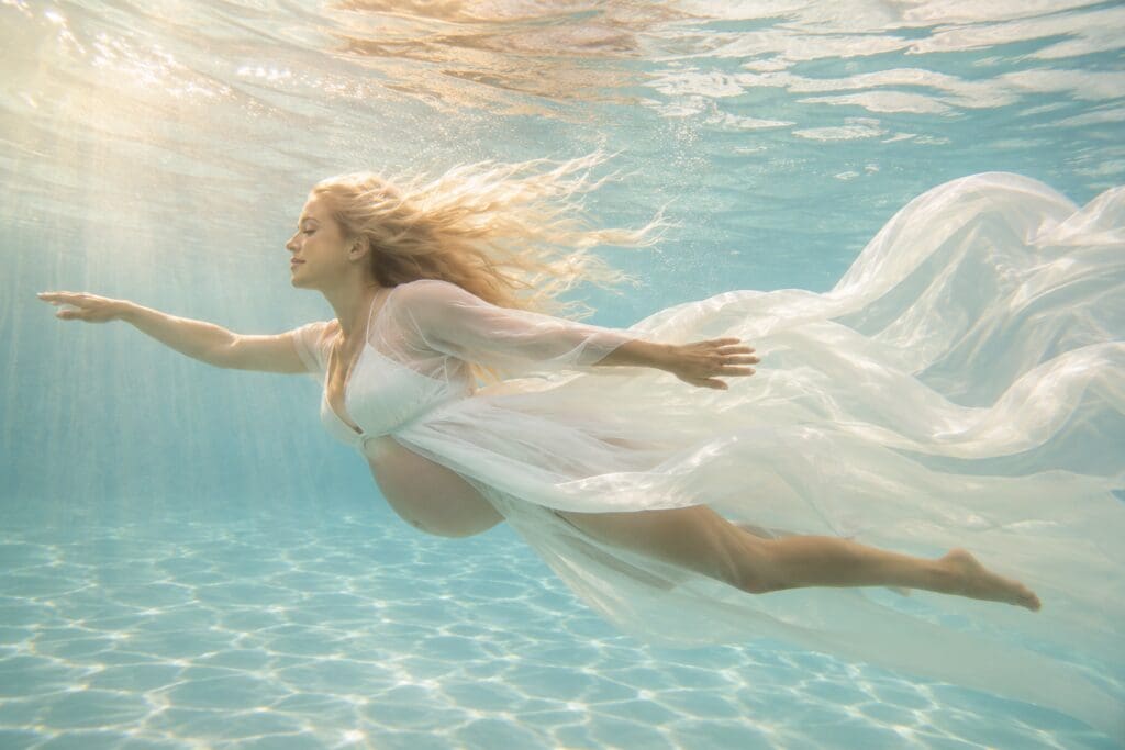 Pregnant woman swimming underwater in a flowing white dress with blonde hair drifting through a pool in Hood River, Oregon