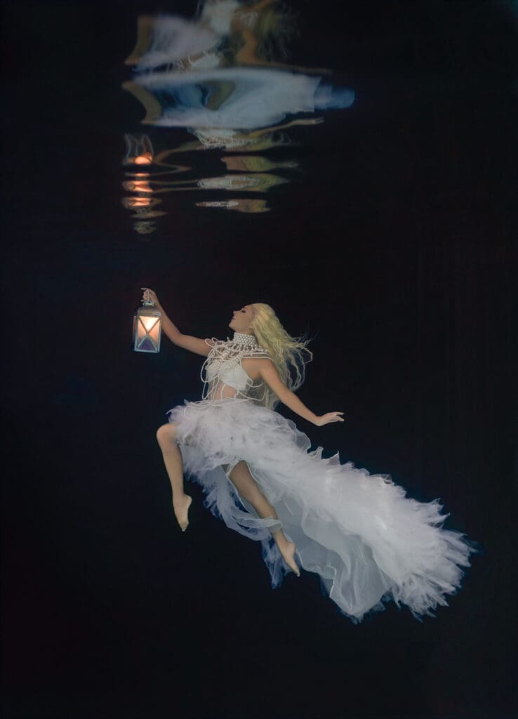 Fine art underwater portrait of Hannah Fraser, known as Hannah Mermaid, in a flowing white dress holding a glowing lantern, photographed by Tobi Rose Rabaliati